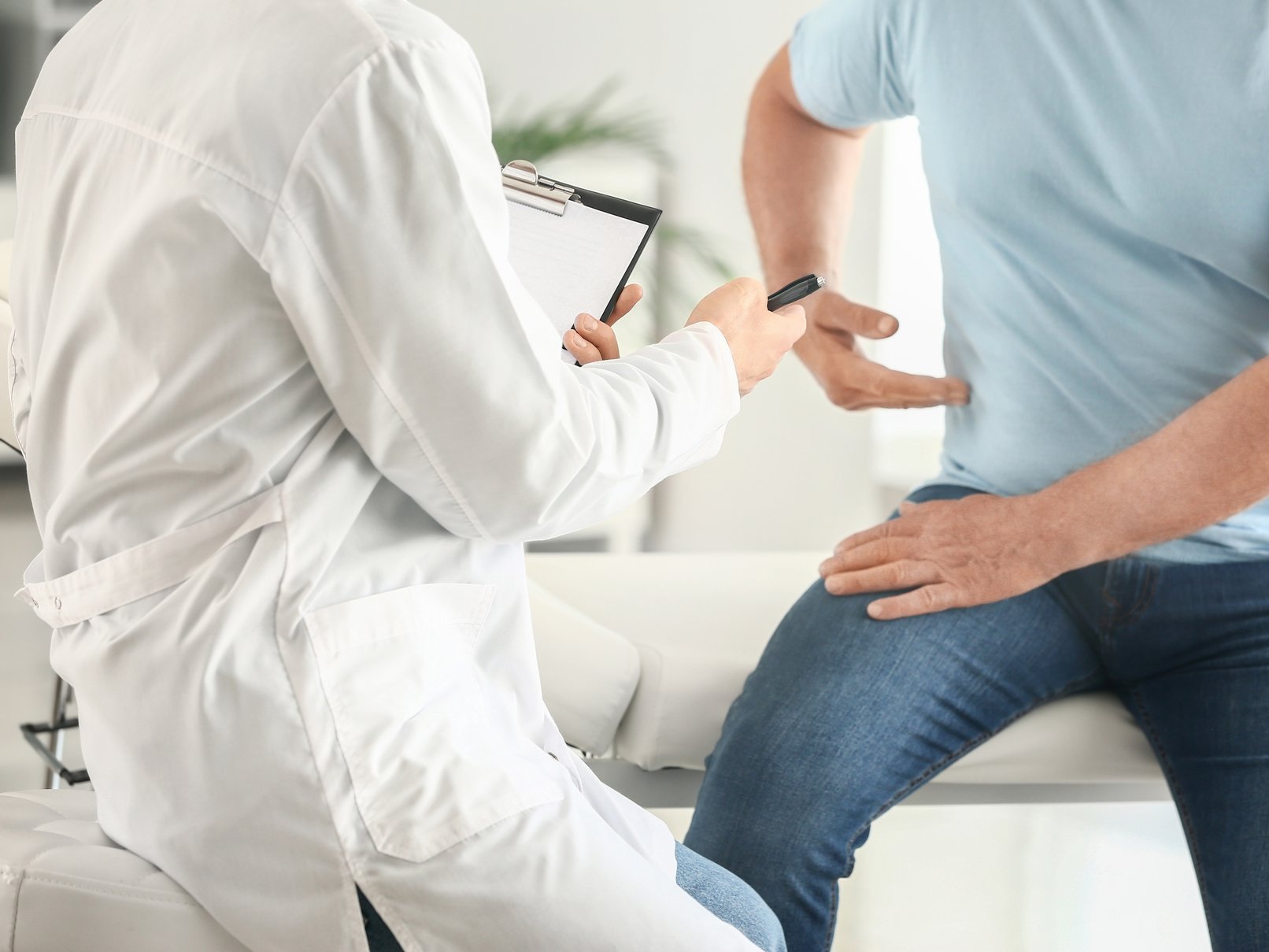 Arzt untersucht einen Patienten mit Rückenschmerzen, der auf der Liege sitzt und auf seine Seite zeigt.